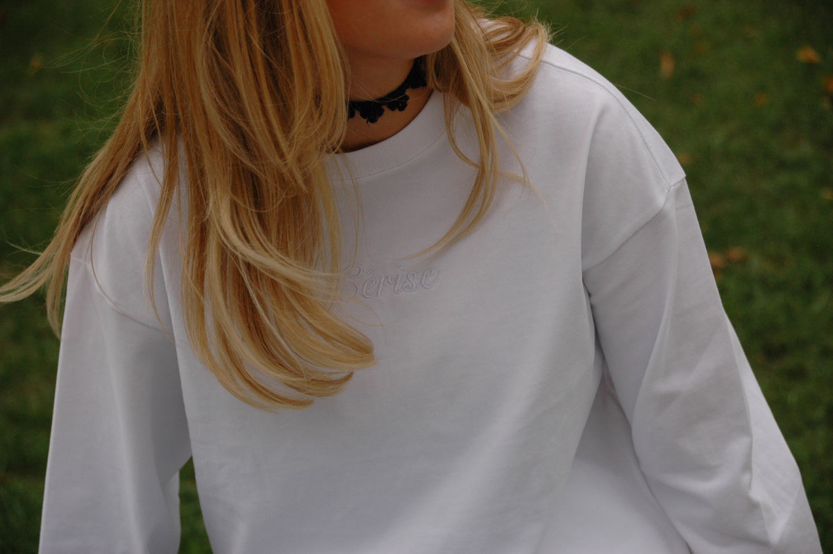 Weißes Cérise Langarmshirt Blanc mit Ton-in-Ton-Schriftzug am Ausschnitt, Model mit blondem Haar und schwarzem Choker vor Wiese