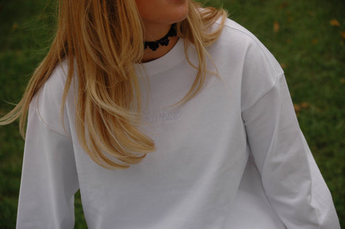 Weißes Cérise Langarmshirt Blanc mit Ton-in-Ton-Schriftzug am Ausschnitt, Model mit blondem Haar und schwarzem Choker vor Wiese