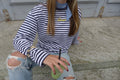Model im blau-weiß gestreiften Langarmshirt O-Three mit gelbem Cérise-Schriftzug, sitzend mit grünem Getränk, Bandana und zerrissener Jeans