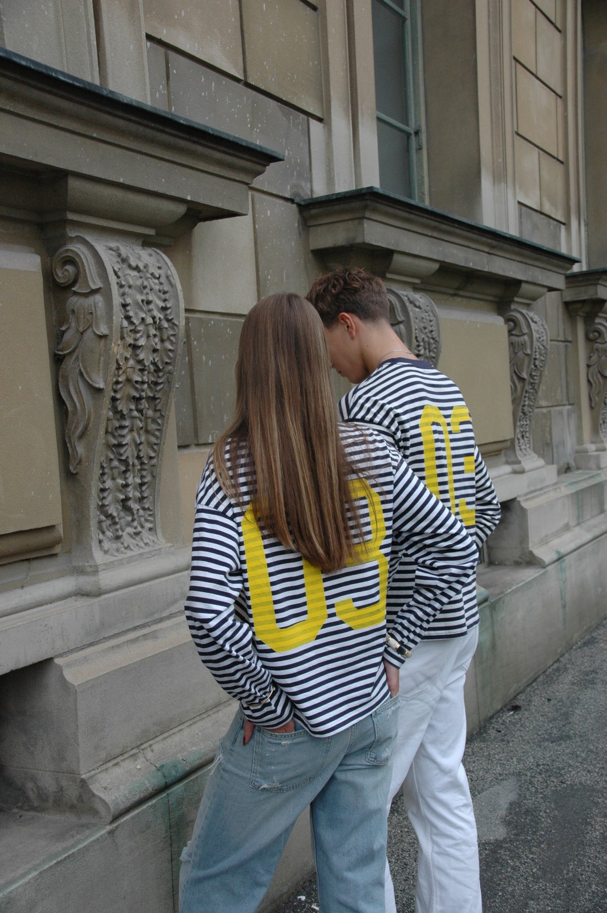 Zwei Models von hinten vor einer Steinfassade in blau-weiß gestreiften Langarmshirts mit großer gelber 03 auf dem Rücken